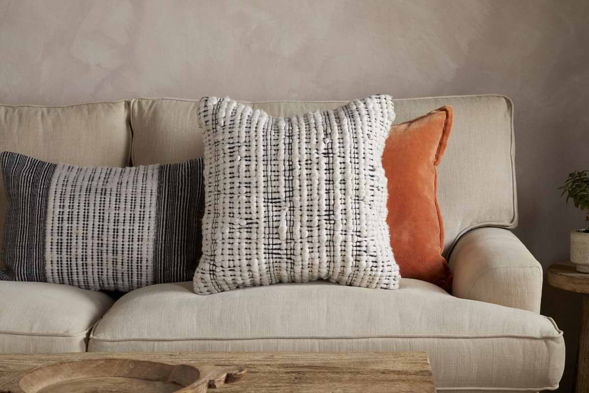 Three decorative pillows rest on a beige sofa: a large textured white-and-black pillow with cushion inners in the center, an nkuku Mika Recycled Cushion Cover - Square in soft orange on the right, and a smaller woven black-and-white pillow crafted from recycled materials on the left. A rustic wooden coffee table sits in front of the sofa.