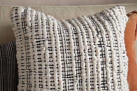 The Mika Recycled Cushion Cover - Square by nkuku features a textured, woven cream fabric with raised, chunky yarn and delicate black grid lines. Styled here on a beige sofa, it’s paired with an assortment of cushions in different colors and textures.