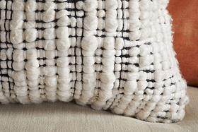 Close-up of a textured, cream-colored Mika Recycled Cushion Cover - Square by nkuku, featuring a chunky woven wool pattern with black thread accents. Made from recycled materials, the cushion cover rests on a beige surface, with a rust-colored pillow partially visible in the background.