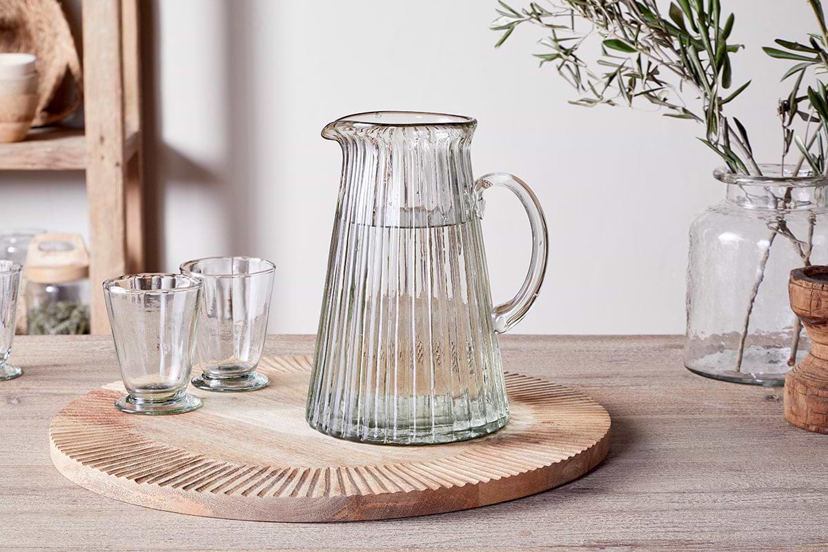 A Minati Ribbed Glass Jug - Clear by nkuku, filled with water, rests on a round wooden tray alongside two empty glasses. Nearby, a glass jar containing olive branches and several wooden kitchen items are artfully arranged on the light-toned tabletop. This scene from the Minati collection creates a rustic, cozy atmosphere with an organic aesthetic.