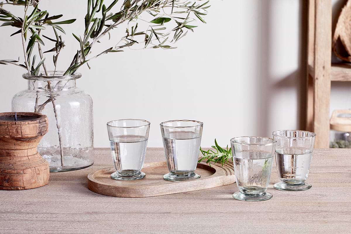 Four clear Minati Tumblers by nkuku, crafted from recycled glass, are arranged on a wooden table. Two tumblers from the set of four rest on an oval wooden tray while the other two sit beside it, bringing a touch of elegance to any tablescape. To the left, a large glass jar filled with leafy branches and a wooden object complement the scene, while a blurred wooden shelf appears in the background.