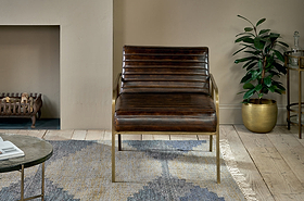 The Mirai Ribbed Leather Armchair by nkuku, featuring horizontal tufting and brass legs, sits on a patterned rug in a living room with light wood floors. A metallic gold potted plant adds warmth, and a fireplace is partially visible nearby.