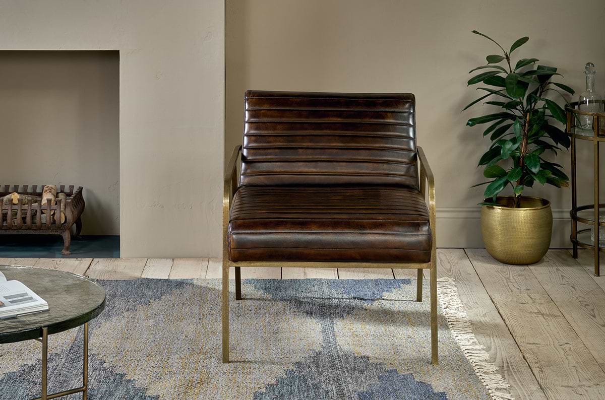The Mirai Ribbed Leather Armchair by nkuku, featuring horizontal tufting and brass legs, sits on a patterned rug in a living room with light wood floors. A metallic gold potted plant adds warmth, and a fireplace is partially visible nearby.