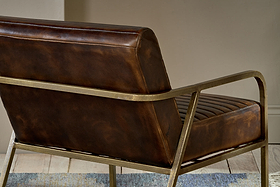 A close-up of the Mirai Ribbed Leather Armchair by nkuku, featuring handmade brown buffalo leather upholstery, ribbed seat detailing, and a sleek gold metal frame, sits on a light wood floor beside a blue-patterned rug.
