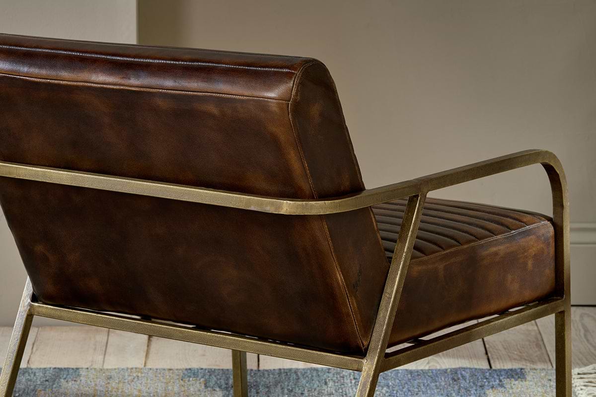 A close-up of the Mirai Ribbed Leather Armchair by nkuku, featuring handmade brown buffalo leather upholstery, ribbed seat detailing, and a sleek gold metal frame, sits on a light wood floor beside a blue-patterned rug.