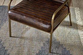 The Mirai Ribbed Leather Armchair by nkuku features modern buffalo leather, horizontal stitching, and slim brass-colored metal legs and armrests. It rests on a textured rug with subtle blue, gray, and beige geometric patterns.