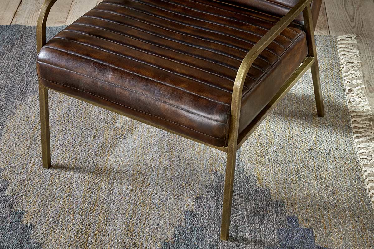 The Mirai Ribbed Leather Armchair by nkuku features modern buffalo leather, horizontal stitching, and slim brass-colored metal legs and armrests. It rests on a textured rug with subtle blue, gray, and beige geometric patterns.