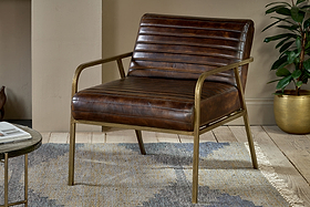 The nkuku Mirai Ribbed Leather Armchair features brown buffalo leather upholstery and slim brass-finished metal arms and legs. It’s styled on a patterned rug with wood floors, beige walls, and a potted plant nearby.