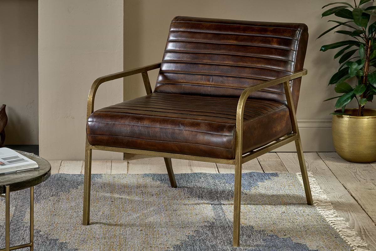 The nkuku Mirai Ribbed Leather Armchair features brown buffalo leather upholstery and slim brass-finished metal arms and legs. It’s styled on a patterned rug with wood floors, beige walls, and a potted plant nearby.