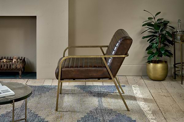 The Mirai Ribbed Leather Armchair by nkuku, crafted from handmade buffalo leather with gold metal arms and legs, stands on a wooden floor near a blue-gray rug. Nearby is a tall green plant in a gold pot against a beige wall, with fireplace and side table in view.