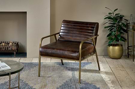 The Mirai Ribbed Leather Armchair by nkuku, upholstered in buffalo leather with cast iron armrests, sits on a patterned rug in a modern living room with a gold planter, fireplace, round side table, wooden floors, and beige walls.