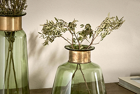 Two Miza Glass Vases - Green by nkuku, each with a decorative brass rim, hold dried branches with pale leaves. The coloured glass vases rest on a wooden surface against a light wall, with part of a closed book visible at the right edge.