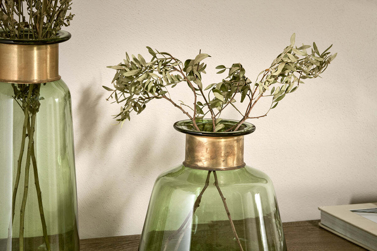 Two Miza Glass Vases - Green by nkuku, each with a decorative brass rim, hold dried branches with pale leaves. The coloured glass vases rest on a wooden surface against a light wall, with part of a closed book visible at the right edge.