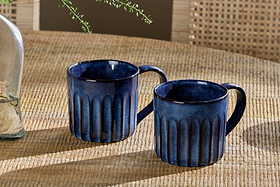 Two nkuku Moda Mugs - Deep Blue (Set of 2) with vertical ribbed patterns rest side by side on a woven beige tabletop, with a glass vase of greenery and a rattan chair back in the background.
