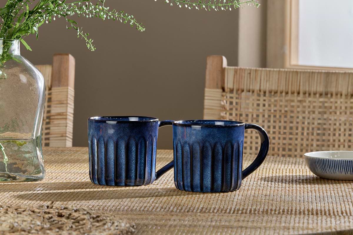 Two Moda Mugs - Deep Blue by nkuku sit side by side on a woven tabletop. In the background, a glass vase with greenery, wicker chairs, and window light create charm, while a small blue-and-white bowl rests on the right.