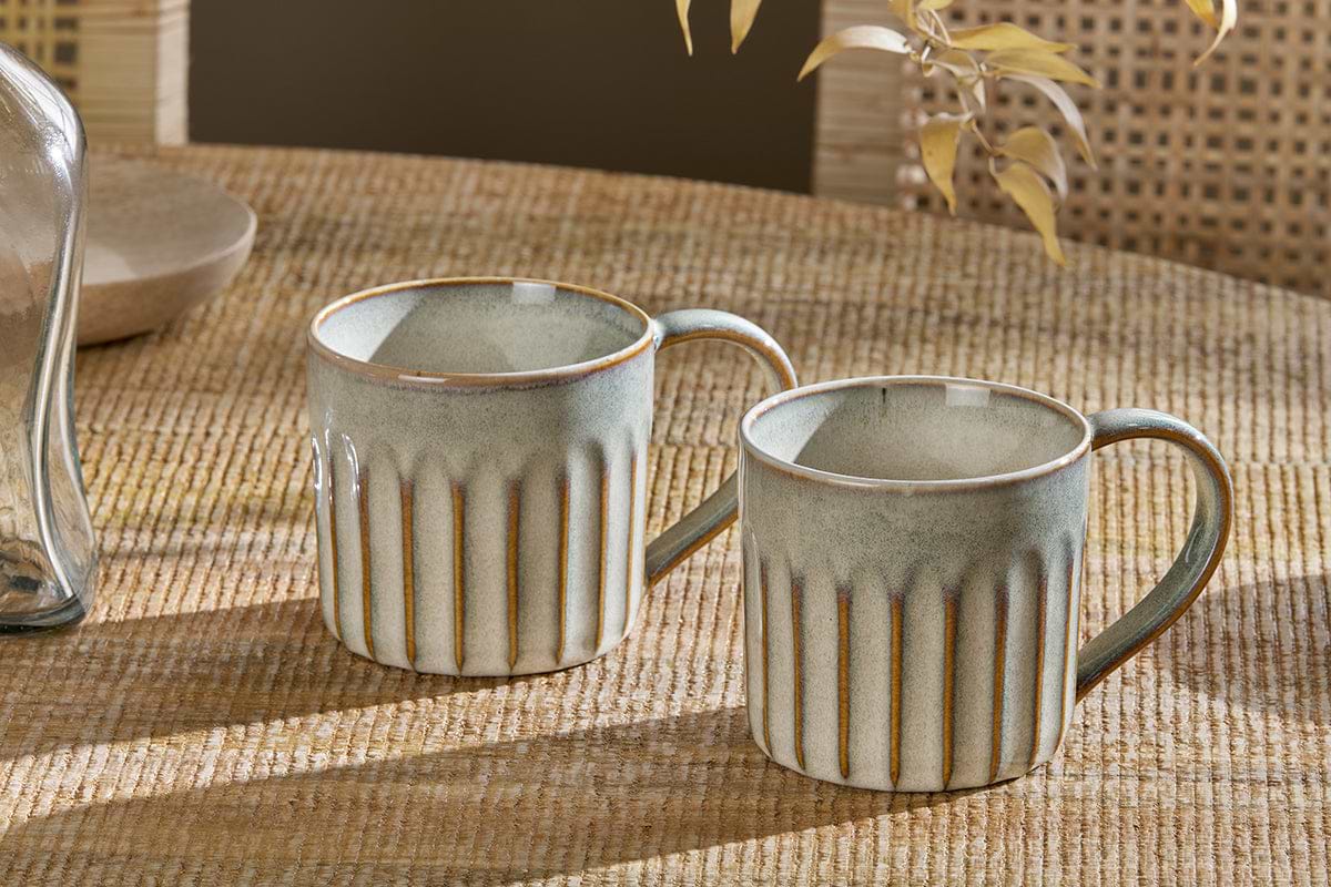 The nkuku Moda Mug - Natural (Set of 2) features ceramic mugs with vertical ridges, a light blue-gray glaze, large handles, and brown-rim accents, displayed on a woven surface with a blurred chair and dried leaves in the background.