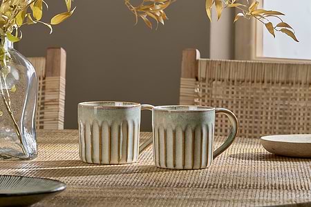 The nkuku Moda Mug - Natural (Set of 2), featuring a ribbed texture and earthy glaze, sits on a woven table. Wooden chairs with cane backs, dried leaves, and warm sunlight create a cozy background.