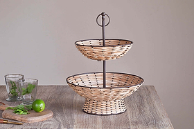The nkuku Montali Rattan 2 Tier Fruit Stand in Black & Natural is displayed on a wooden kitchen counter with two empty glasses, mint sprigs, a lime, and a knife on a small cutting board against a plain light background.