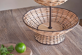 The Montali Rattan 2 Tier Fruit Stand by nkuku sits on a wooden table, with its lower, empty basket in view. Beside it, a wooden board holds a whole lime and mint leaves, adding rustic charm to any kitchen counter.