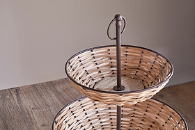 The Montali Rattan 2 Tier Fruit Stand by nkuku, in black & natural with metal accents and a central rod, sits empty on a wooden kitchen counter with light walls that highlight its elegant design.