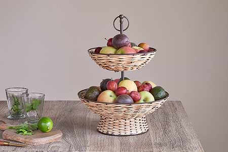 The nkuku Montali Rattan 2 Tier Fruit Stand in Black & Natural sits on a wooden table, filled with apples, plums, avocados, and limes. Nearby are two empty glasses and a cutting board with mint leaves, a lime, and a knife against a plain background.