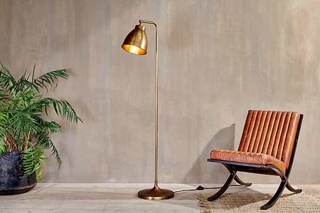 A modern space showcases the nkuku Muturi Floor Lamp - Antique Brass with a sleek down light, angled left beside a brown leather chair. A green plant sits nearby on light wood floors with a woven rug and beige walls.