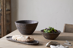 A wooden table set with nkuku’s Nyati Wooden Serving Bowl – Dark Brown – Small, filled with greens, plus a long wooden tray holding bread. A fork, knife, and napkin are placed nearby.
