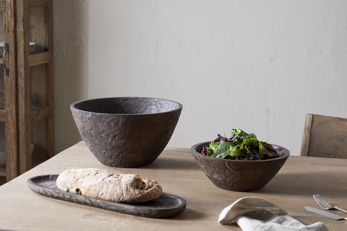 A wooden table set with nkuku’s Nyati Wooden Serving Bowl – Dark Brown – Small, filled with greens, plus a long wooden tray holding bread. A fork, knife, and napkin are placed nearby.