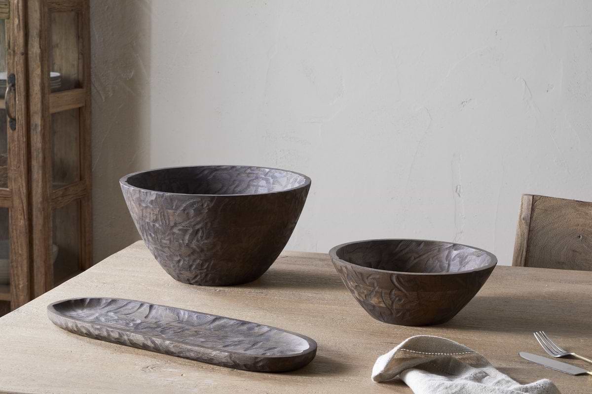 The nkuku Nyati Wooden Serving Bowl in dark brown (small) sits on a wooden table near a folded napkin, fork, and spoon, with a rustic cabinet in the background.
