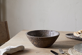 The nkuku Nyati Wooden Serving Bowl in dark brown, small, sits empty on a light wood table with a napkin, fork, and a plate of bread nearby, against a plain off-white wall background.
