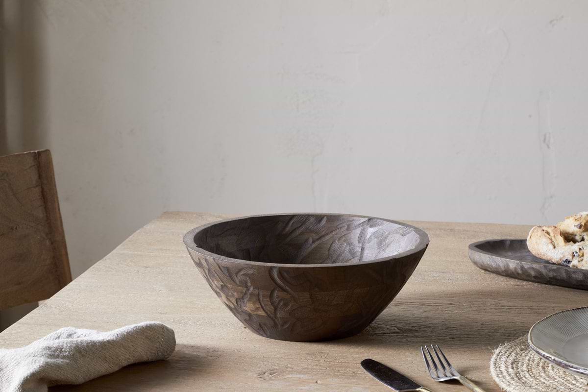 The nkuku Nyati Wooden Serving Bowl in dark brown, small, sits empty on a light wood table with a napkin, fork, and a plate of bread nearby, against a plain off-white wall background.