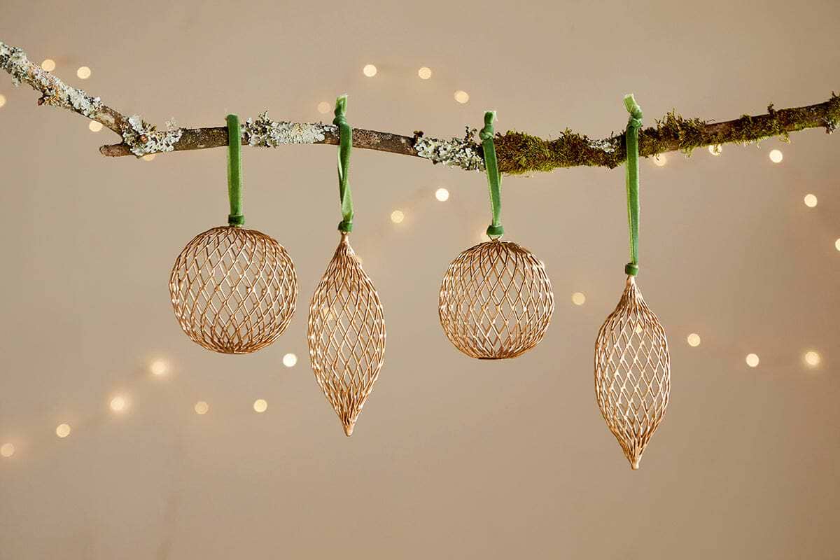The Nirav Wire Baubles - Brass (Set of 4) from nkuku feature round and elongated brass-finished ornaments, each hung by a green ribbon on a mossy branch, softly glimmering against warm white fairy lights.