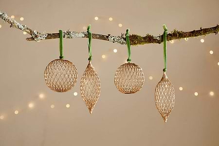 The Nirav Wire Baubles - Brass (Set of 4) from nkuku feature round and elongated brass-finished ornaments, each hung by a green ribbon on a mossy branch, softly glimmering against warm white fairy lights.