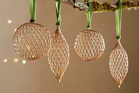The Nirav Wire Baubles - Brass (Set of 4) by nkuku, featuring three spherical and one elongated bauble, hang from a moss-accented branch with green ribbon. Warm bokeh lights in the background create an elegant festive display.