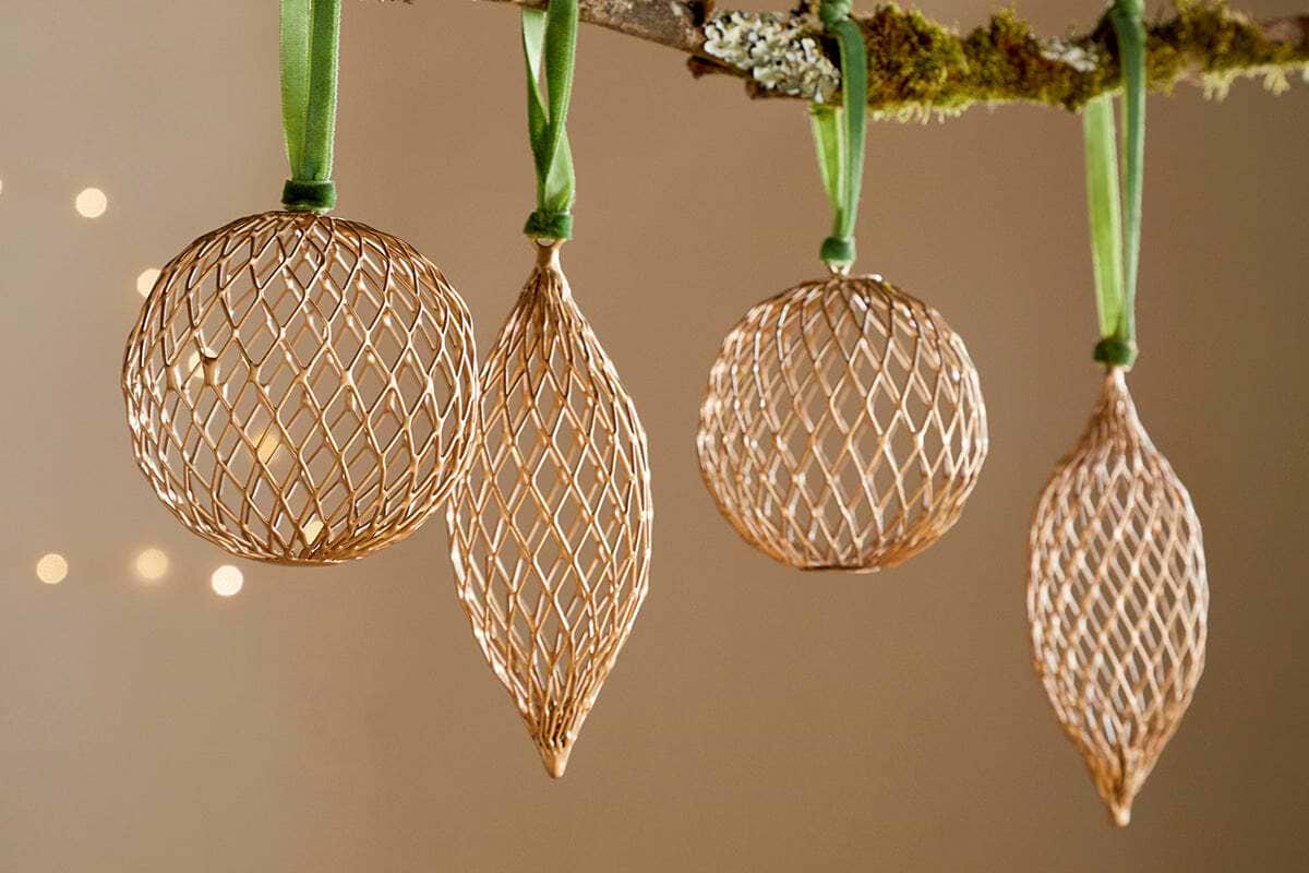 The Nirav Wire Baubles - Brass (Set of 4) by nkuku, featuring three spherical and one elongated bauble, hang from a moss-accented branch with green ribbon. Warm bokeh lights in the background create an elegant festive display.