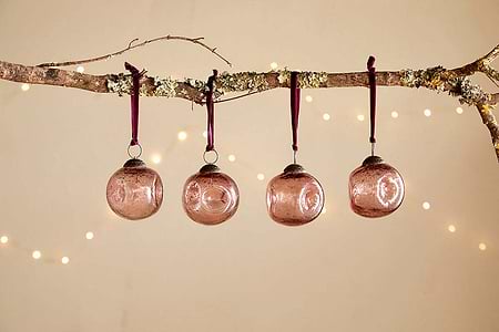 A bare branch displays four nkuku Nishan Pinched Baubles in copper, each hung with a burgundy velvet tie. Warm, blurred lights glow softly against a beige backdrop, adding festive charm to the recycled glass decorations.