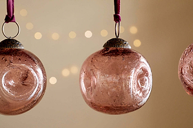 Four copper Nishan Pinched Baubles by nkuku, featuring a unique pinched texture and silver caps, hang from velvet ties. Set against a soft beige background with blurred festive lights creating a warm ambiance.