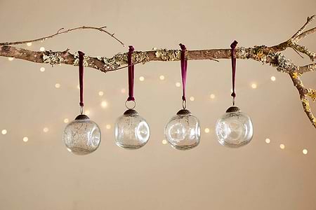 A bare branch with moss and lichen displays four nkuku Nishan Pinched Baubles - Clear, each hung by burgundy velvet ties. Soft fairy lights glow in the background, creating an elegant and festive beige-wall display.