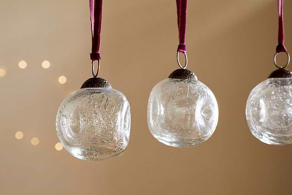 Four clear recycled glass Nishan Pinched Baubles by nkuku hang from burgundy velvet ties against a neutral beige backdrop, with soft, out-of-focus lights creating a warm, festive glow.
