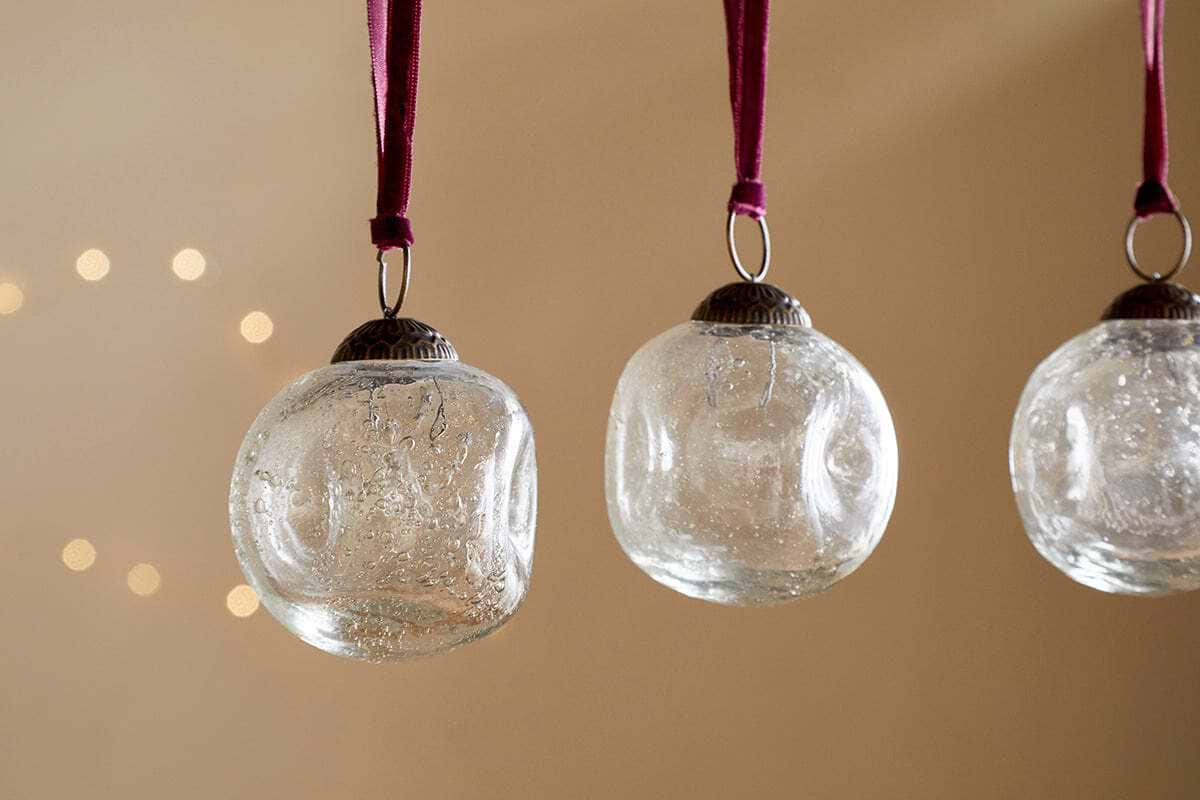 Four clear recycled glass Nishan Pinched Baubles by nkuku hang from burgundy velvet ties against a neutral beige backdrop, with soft, out-of-focus lights creating a warm, festive glow.