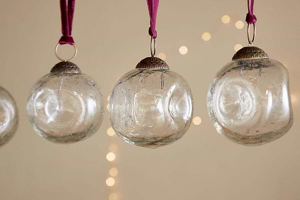 The nkuku Nishan Pinched Baubles - Clear (Set of 4) feature gently irregular shapes and hang from burgundy velvet ties, their recycled glass shimmering softly against a neutral backdrop with twinkling string lights for a festive touch.