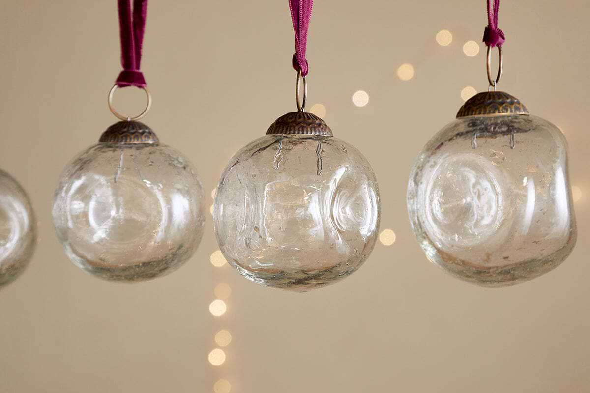 The nkuku Nishan Pinched Baubles - Clear (Set of 4) feature gently irregular shapes and hang from burgundy velvet ties, their recycled glass shimmering softly against a neutral backdrop with twinkling string lights for a festive touch.