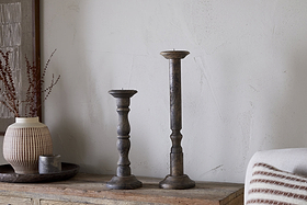 Two Nayati Rustic Wood Pillar Candle Sticks by nkuku in dark stain, each of different heights, stand on a wooden surface beside a ribbed ceramic vase with dried stems and a small metal container on a tray against an off-white textured wall.