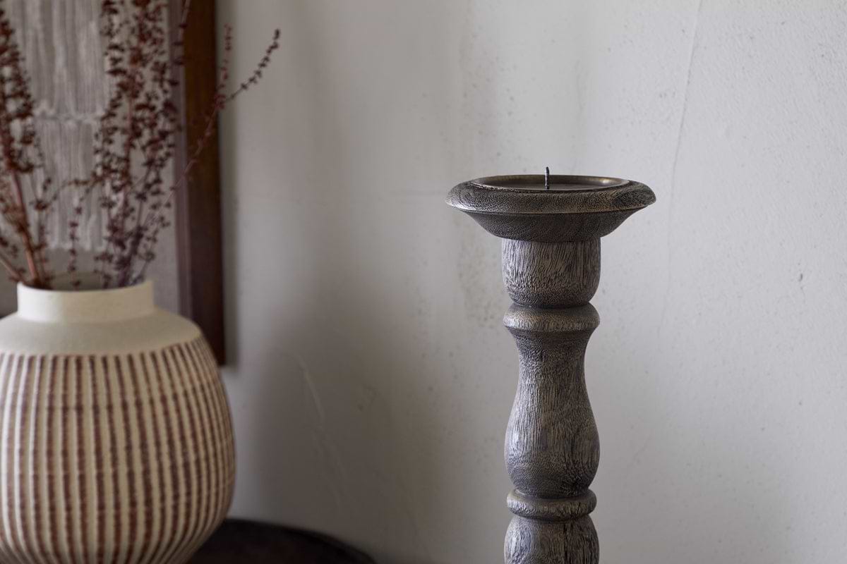 The Nayati Rustic Wood Pillar Candle Stick by nkuku, in a dark stain, stands beside a beige vase with brown stripes and dried branches. A white textured wall behind them adds to the cozy, minimal decor.