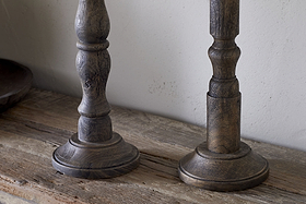 The Nayati Rustic Wood Pillar Candle Stick by nkuku features turned detailing and a dark stain, crafted from sustainable mango wood. Its visible grain and worn texture enhance its vintage charm against a plain, light-colored wall.