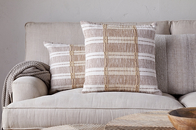 Two Nayika Cotton Cushion Covers by nkuku in rust add warmth to a light tan sofa, with a knitted throw over the armrest. The neutral setting and cosy decor create an inviting atmosphere.