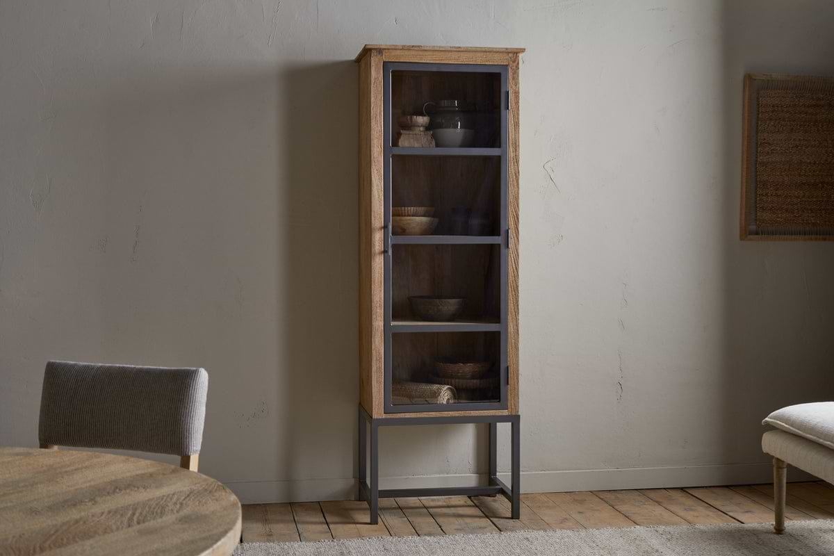 The nkuku Nirvi Mango Wood & Iron Tall Cabinet - Black features glass doors, metal legs, and shelves with bowls and pottery, set against a light wall in a minimalist room with wooden floors and simple furnishings.