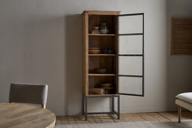 Against a light wall stands the Nirvi Mango Wood & Iron Tall Cabinet by nkuku, with a glass door and shelves showing bowls, plates, and vases. Nearby are a round wooden table, an upholstered chair, and part of a woven wall hanging.