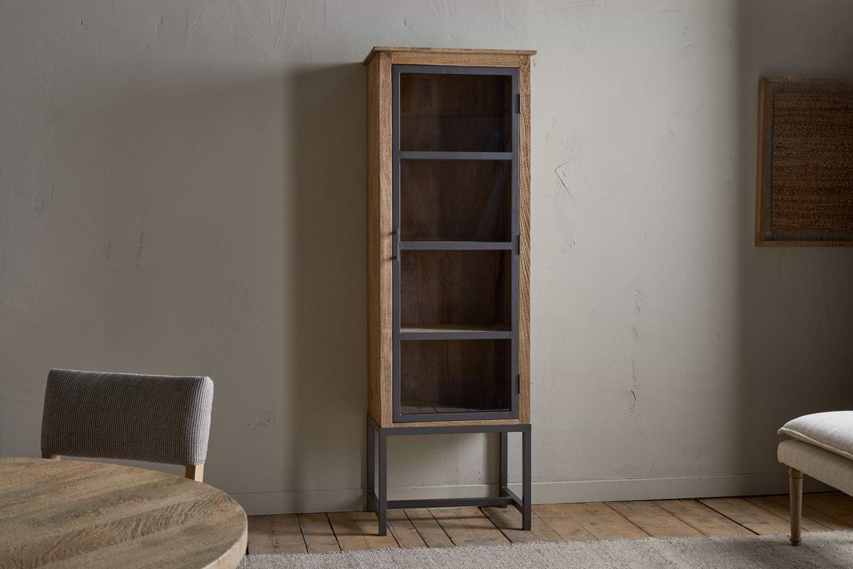 The nkuku Nirvi Mango Wood & Iron Tall Cabinet - Black is a slim, tall display unit with a glass door and metal frame, shown on a wooden floor beside a dining table in a minimalist room with light-colored walls.