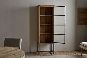 The Nirvi Mango Wood & Iron Tall Cabinet by nkuku, featuring four shelves and a glass door on metal hinges, stands against a light wall. The door is open, with a round wooden table and cushioned chair partially visible in the foreground.